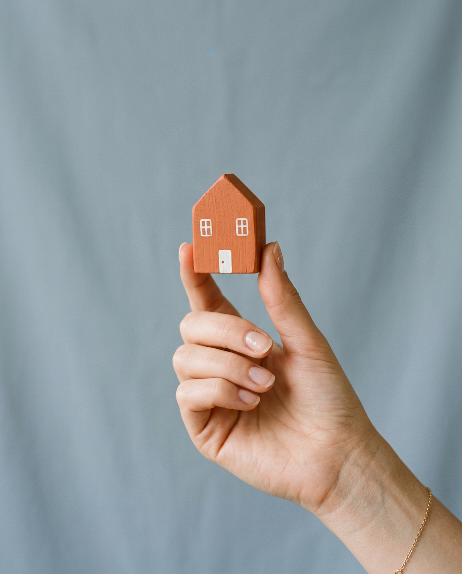 Une main tient une petite maison en bois terracotta devant un fond bleu-gris